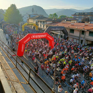 Sportful-Dolomiti-Race - Tour de Vacance Wielervakanties Hundreds of cyclists line a town street at the race start under a red Enervit arch, with a yellow Ricola arch and cheering spectators nearby.