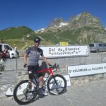 Q8zB6GnF1gBvLDPxXEmt2kHs0ixRJZqRcqVPmYw1SKw-2048x1536jpg - Tour de Vacance Wielervakanties Cyclist in helmet and sunglasses with a red-and-black bike, in front of a stone sign for Col du Glandon at 1924 m, with a alpine backdrop.