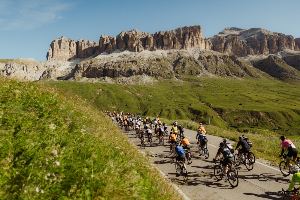 A large group of cyclists ride along a mountain road under a blue sky, with green meadows and rocky cliffs in the background, delivering a scenic cycling scene.