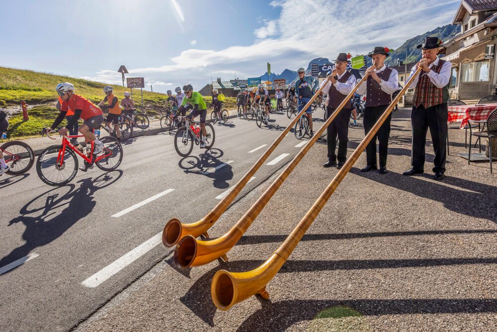 Cyclists ride along a mountain road as four men in traditional attire play long alphorns in the foreground.