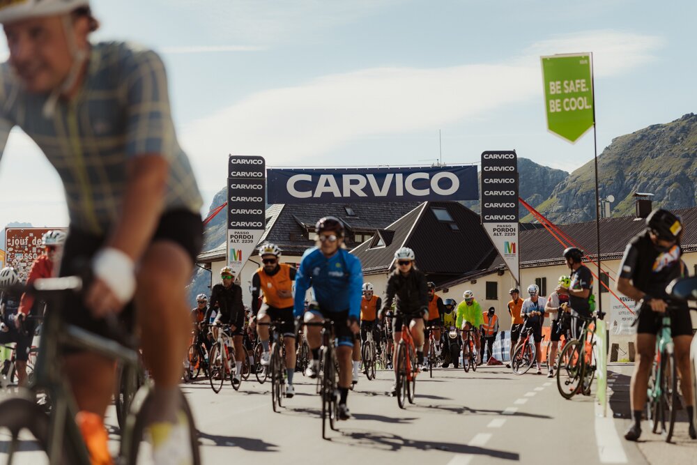 Group of cyclists riding on a city street during a race, with a large CARVICO banner and tents in the background.