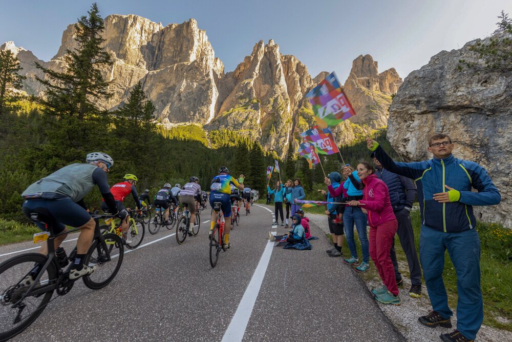Group of cyclists racing along a mountain road as spectators cheer with colorful flags beside towering rock cliffs.