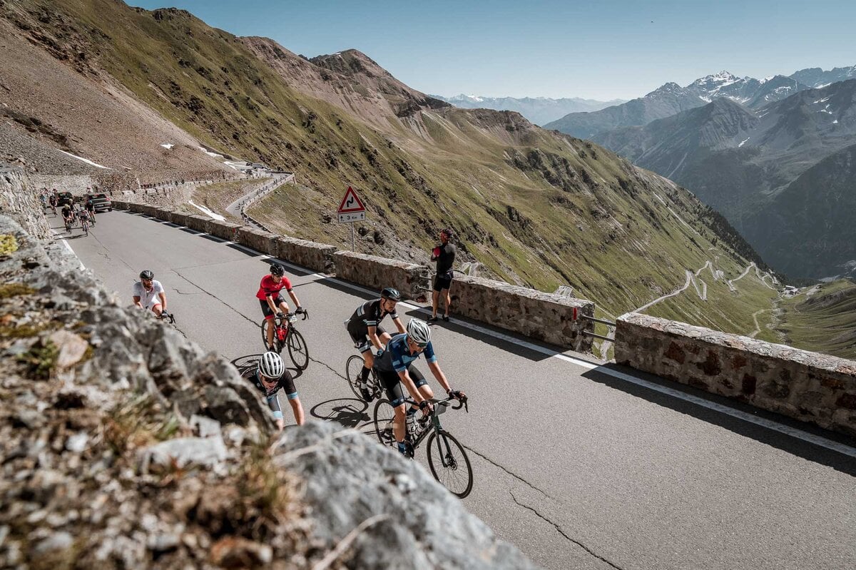 Granfondo Arrangementen - Tour de Vacance Wielervakanties Group of cyclists riding uphill on a narrow mountain road with stone barriers and winding switchbacks amid green slopes and distant snow-capped peaks.