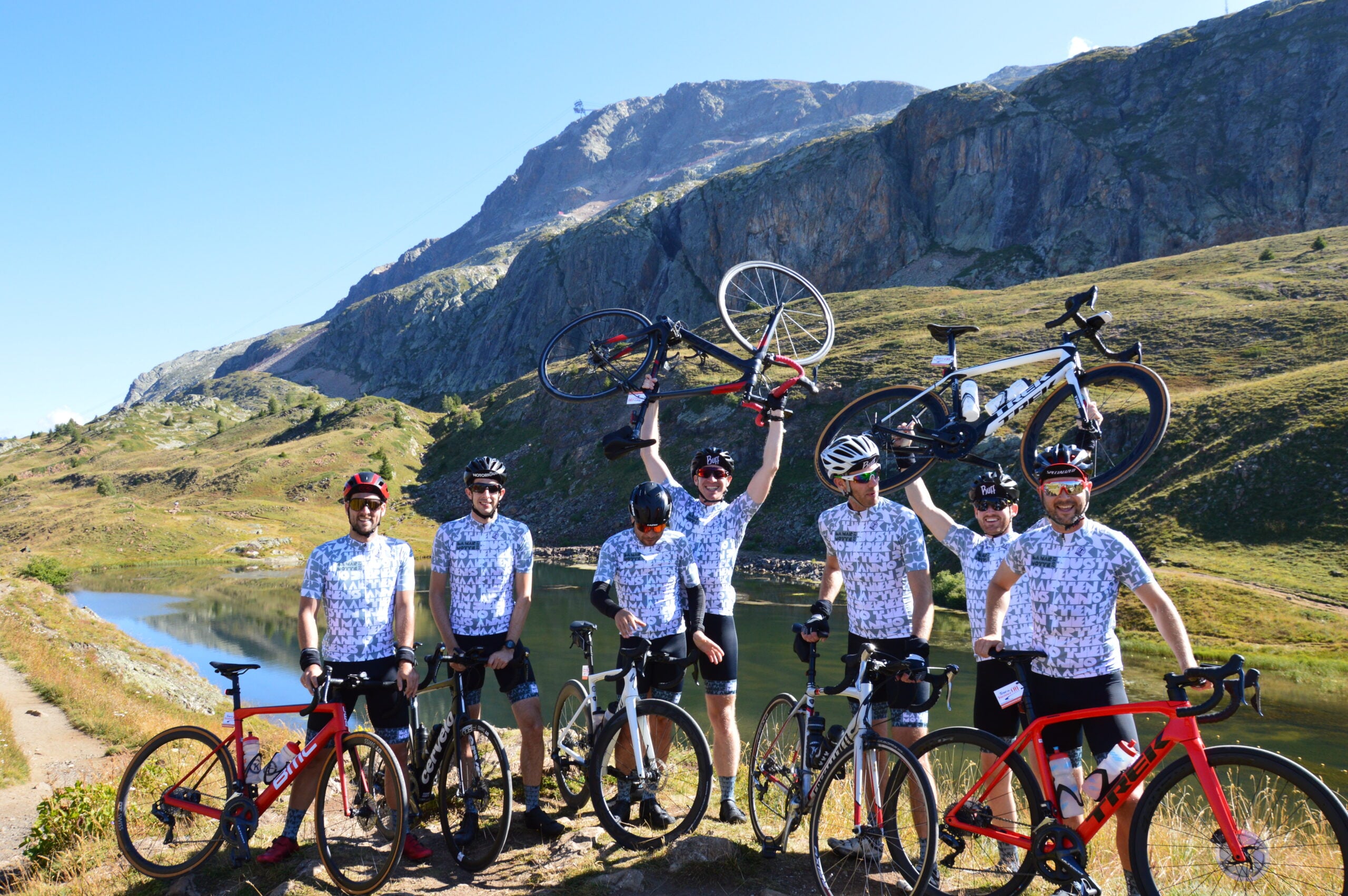 Fietsarrangementen - Tour de Vacance Wielervakanties Group of cyclists in matching jerseys lift their bikes in a mountainous area with a clear blue sky behind them.