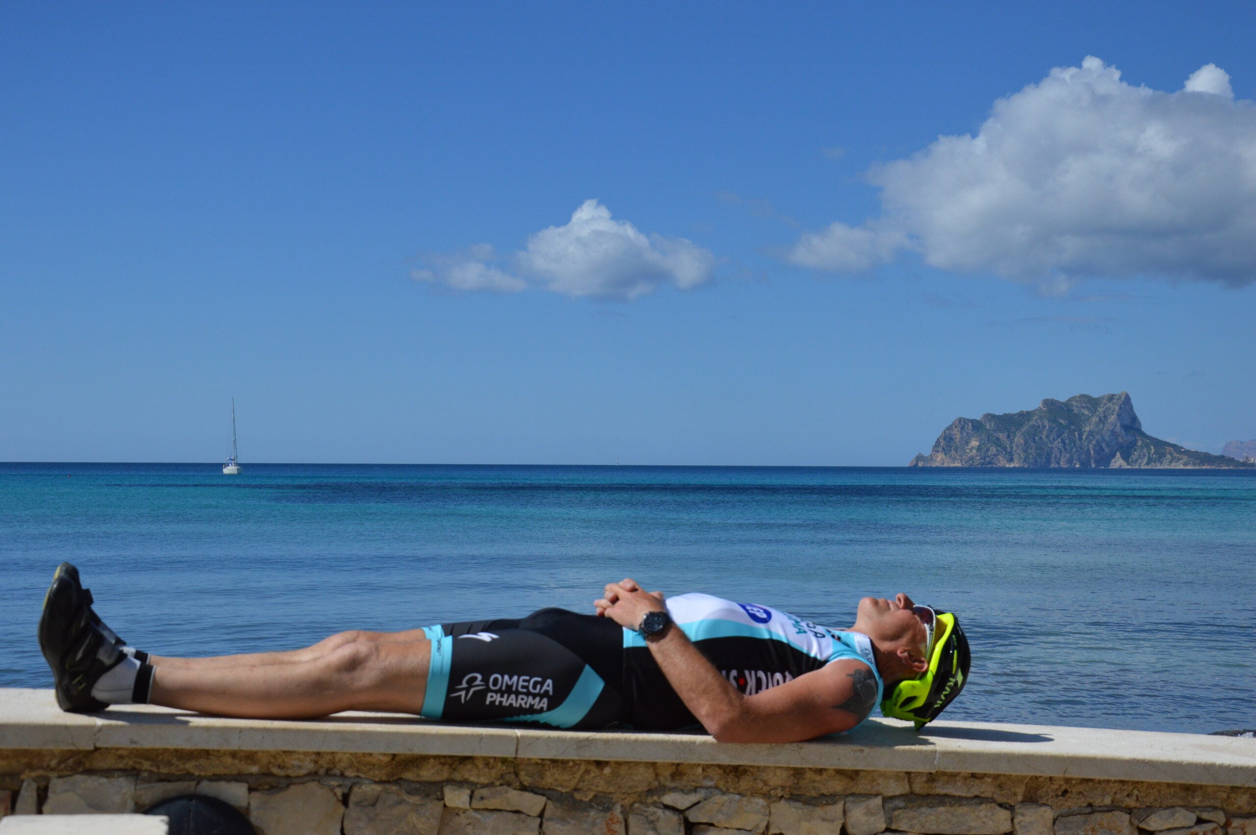 Costa Blanca - Tour de Vacance Wielervakanties Person wearing cycling gear and helmet lying on a seawall with arms crossed, resting by the turquoise sea and distant hills/rocks on the horizon.