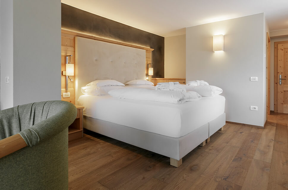 Hotel room with a large white bed, tufted headboard, wooden floor, and warm bedside lamps; a green chair is in the foreground.