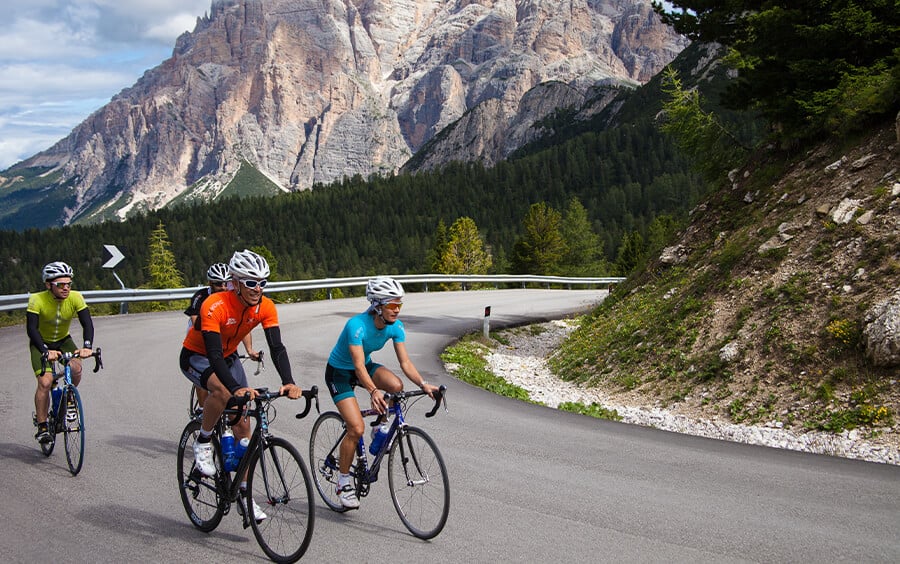 Four cyclists wearing helmets ride along a winding mountain road with rocky cliffs and evergreen forest in the background, under a partly cloudy sky.