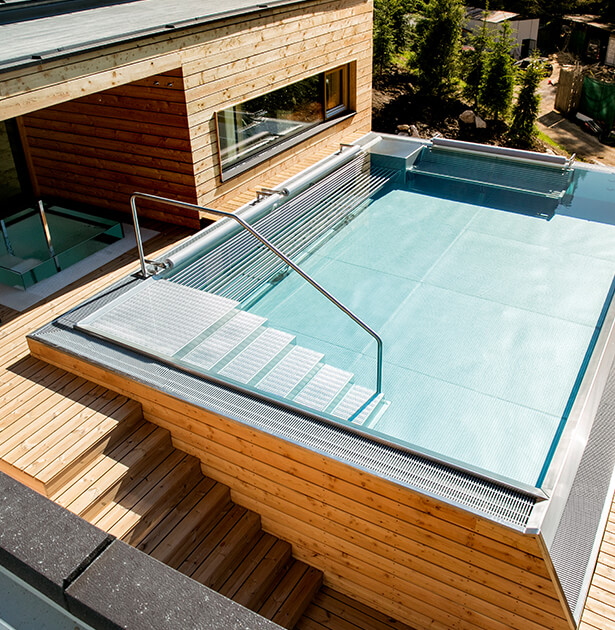 Rectangular outdoor pool on a wooden deck, with a metal handrail and white steps descending to the water level near a modern house window.