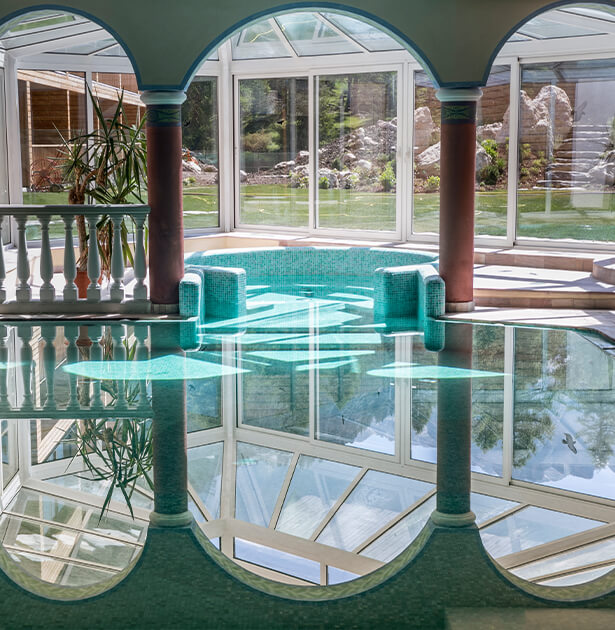 Indoor pool inside a glass-walled room with teal edging, arched green ceiling, and red columns; water reflects the surrounding architecture.