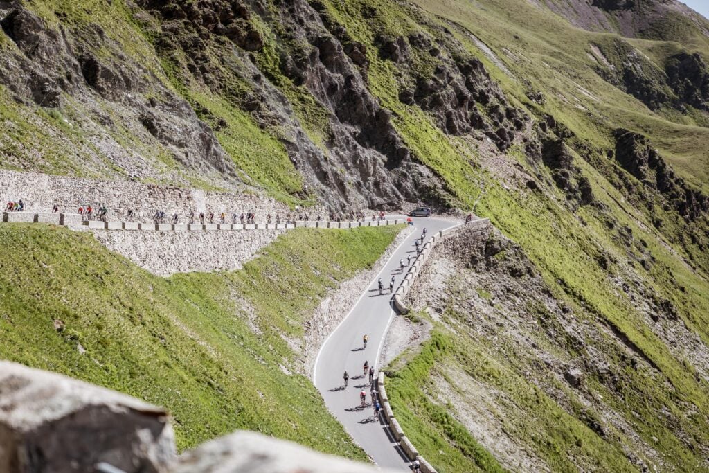 Dreilandergiro - Tour de Vacance Wielervakanties Cyclists ride along a winding mountain road with stone barriers, flanked by steep green cliffs and rock faces.